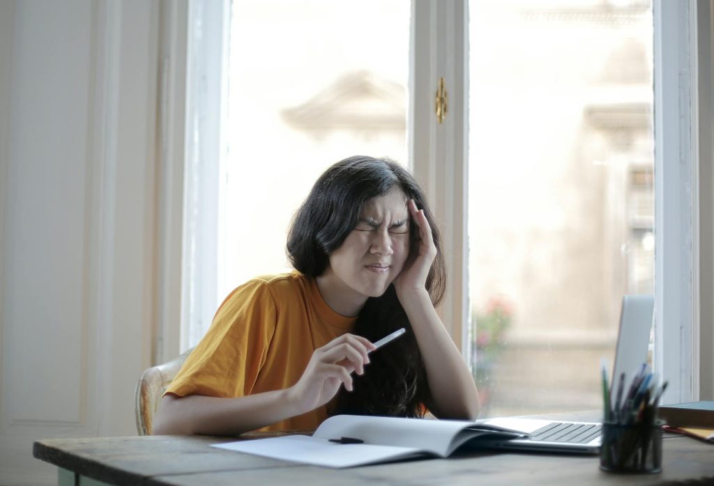 Ethnic female student in casual wear sitting at table with laptop and notebook and grimacing from migraine while doing homework during distance learning